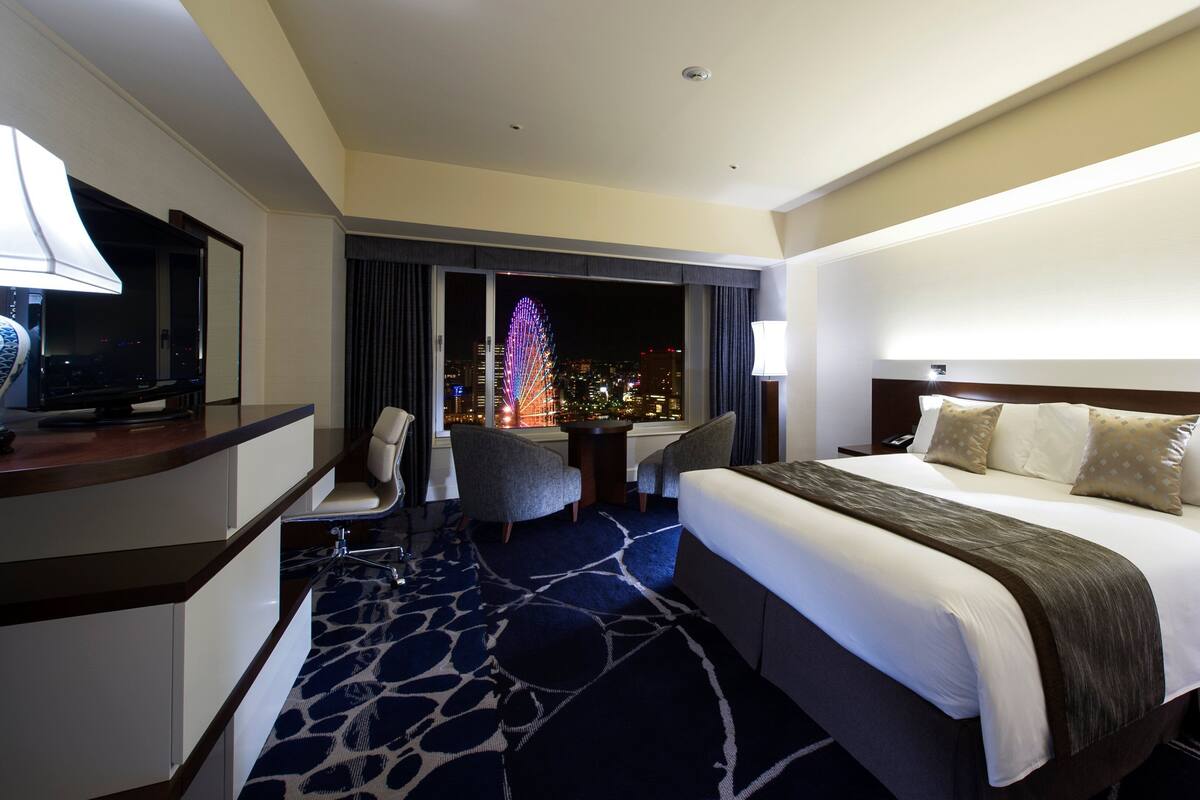 A hotel room with a large bed, a desk with a TV, a chair, and a view of a cityscape with a lit-up Christmas tree.