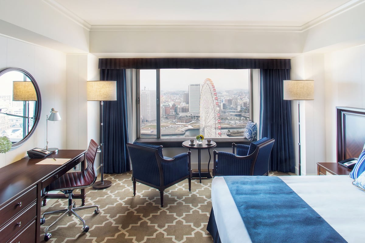 A hotel room with a bed, desk, chair, and a large window offering a city view.