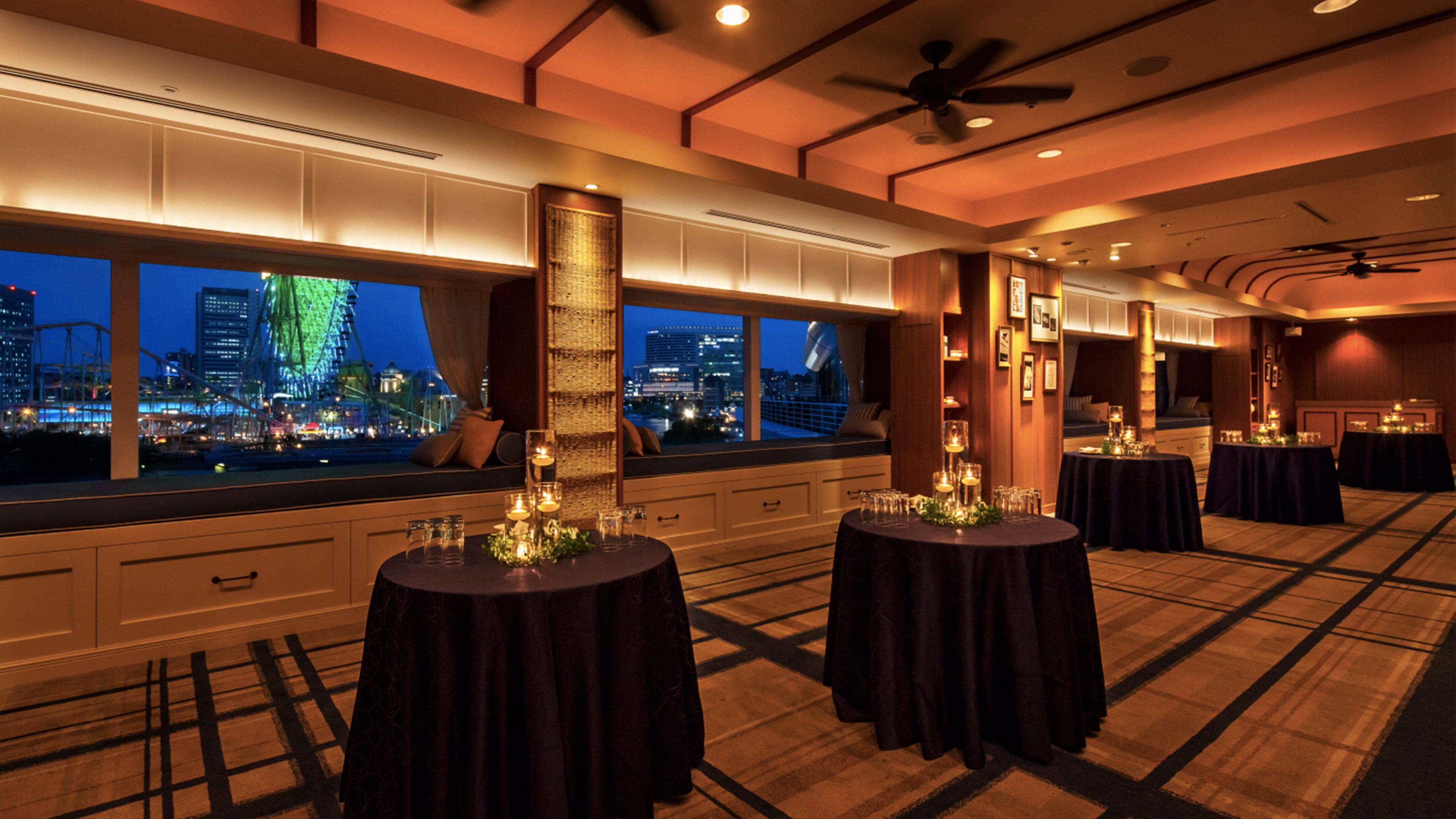 A hotel room with a city view, large windows, and multiple round tables set up for dining.