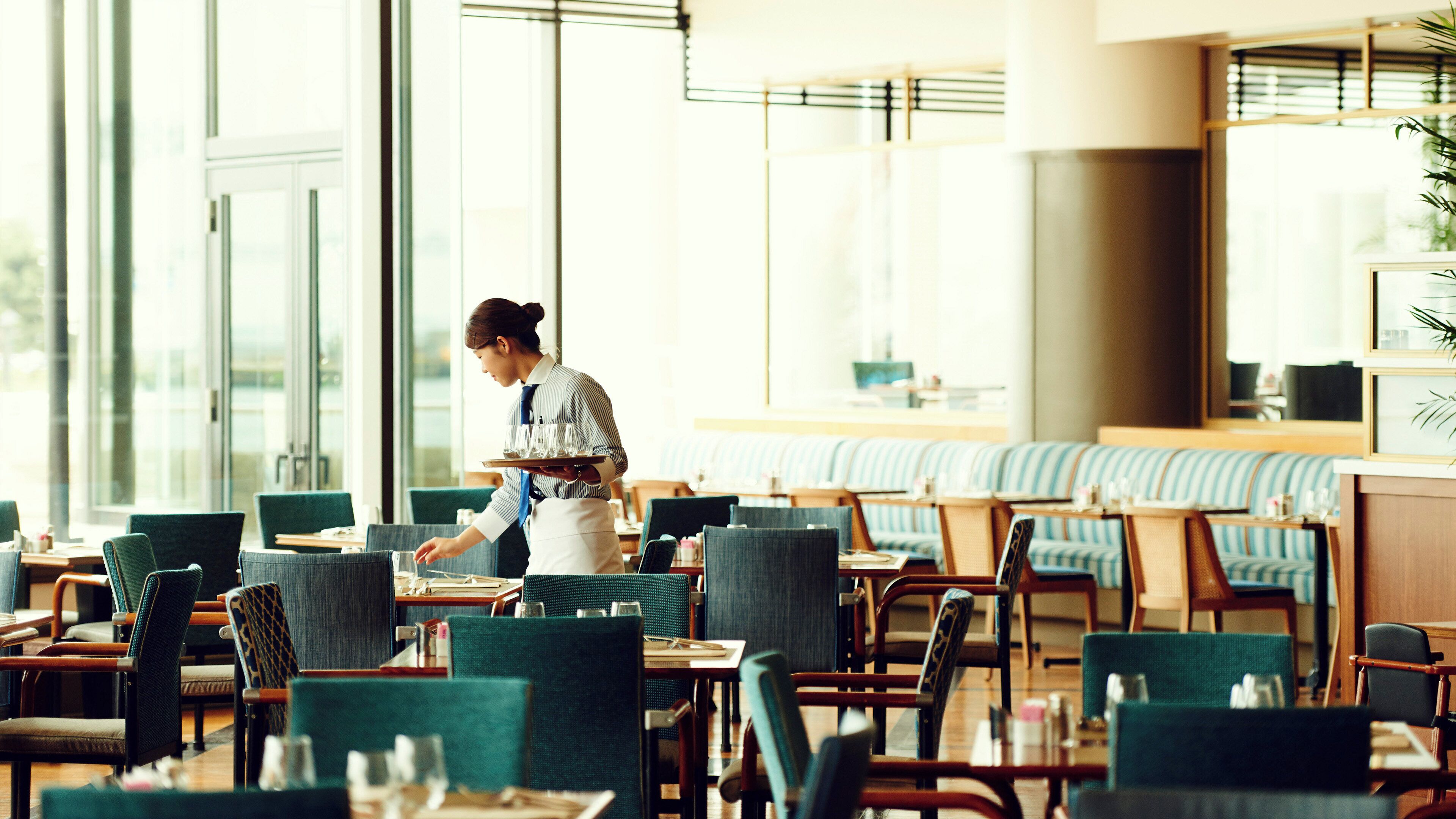 Une salle à manger d’hôtel avec des tables et des chaises, un serveur portant un plateau, et de grandes fenêtres laissant entrer la lumière naturelle.