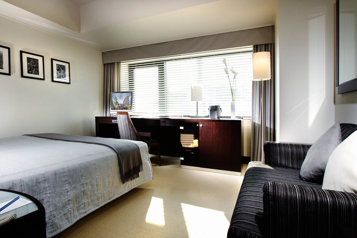 A hotel room with a bed, a desk with a computer, a chair, a sofa, and a window with blinds.