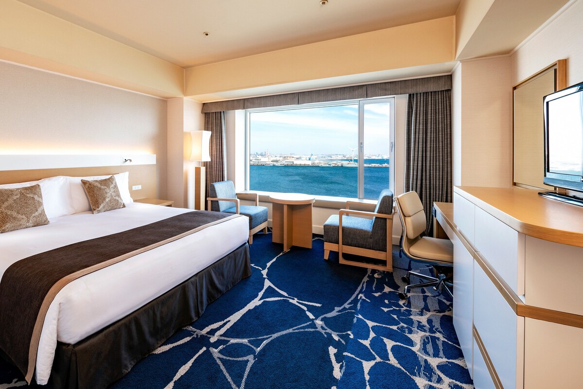 A hotel room with a large bed, a desk, a chair, a TV, and a window with a view of the sea.
