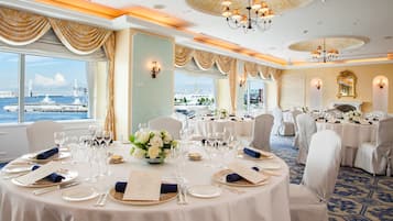 A formal dining area with round tables set for a meal, a view of the sea, and a chandelier.
