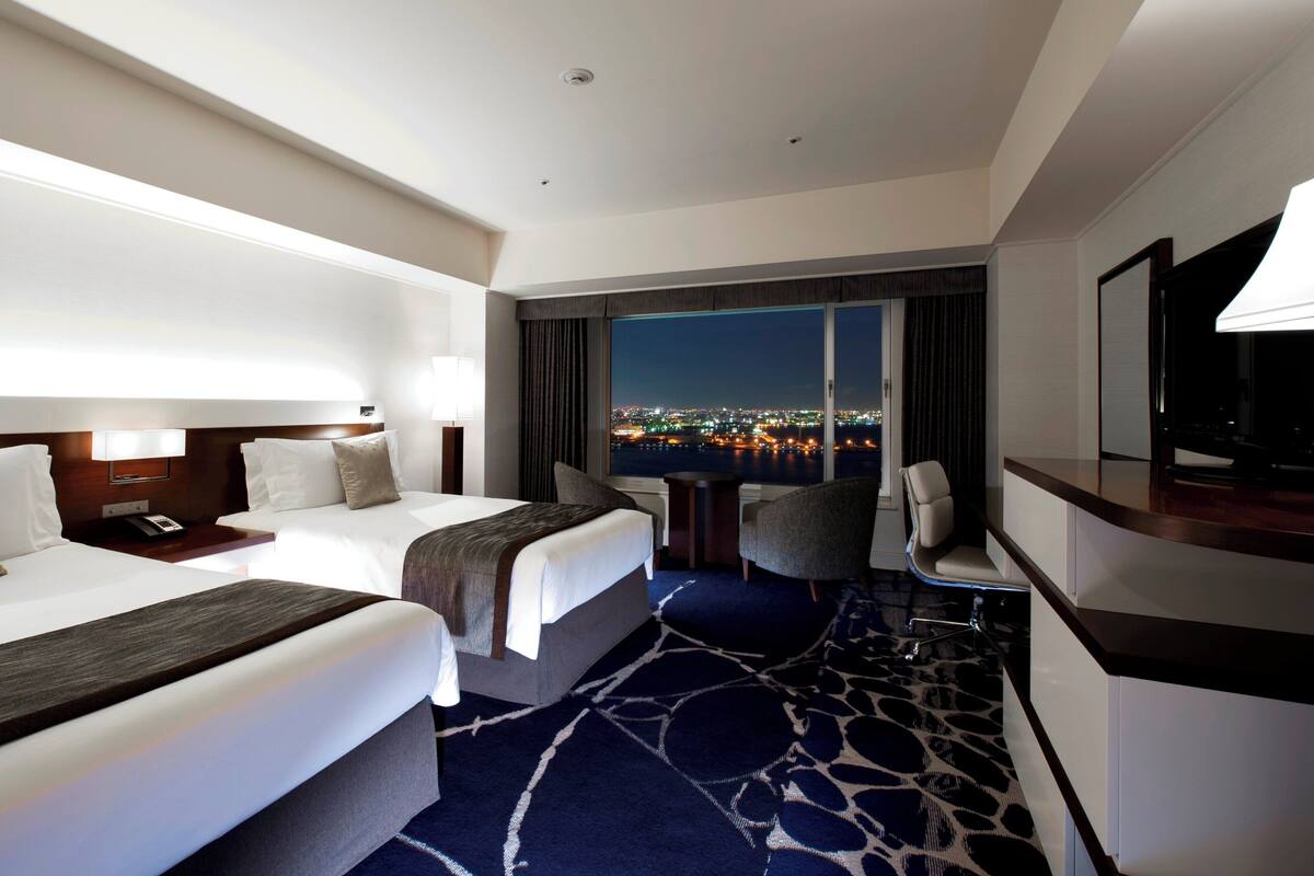 A hotel room with two beds, a desk, a chair, a TV, and a large window with a city view.