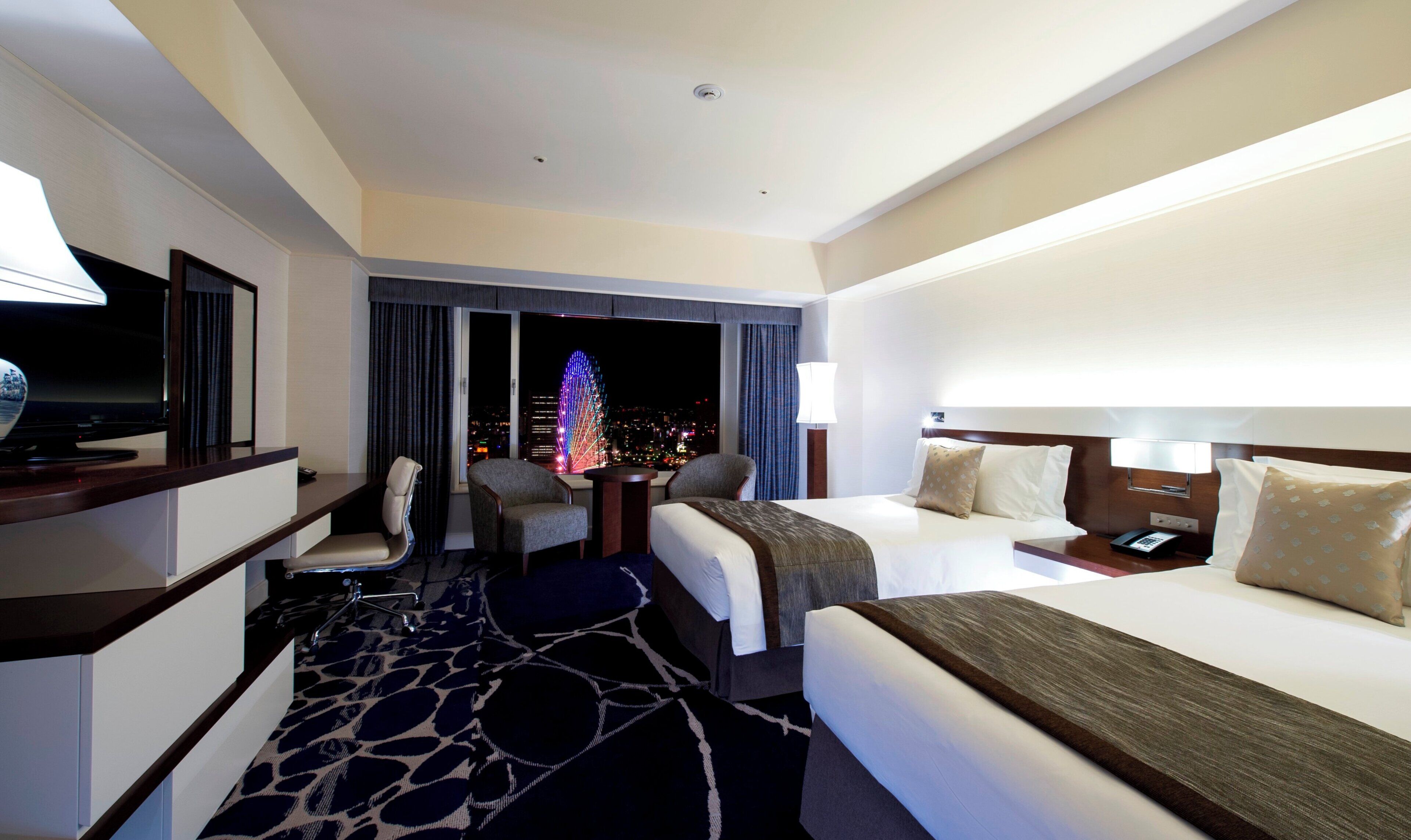 A hotel room with a bed, a desk with a television, a chair, a nightstand, and a view of the city at night.