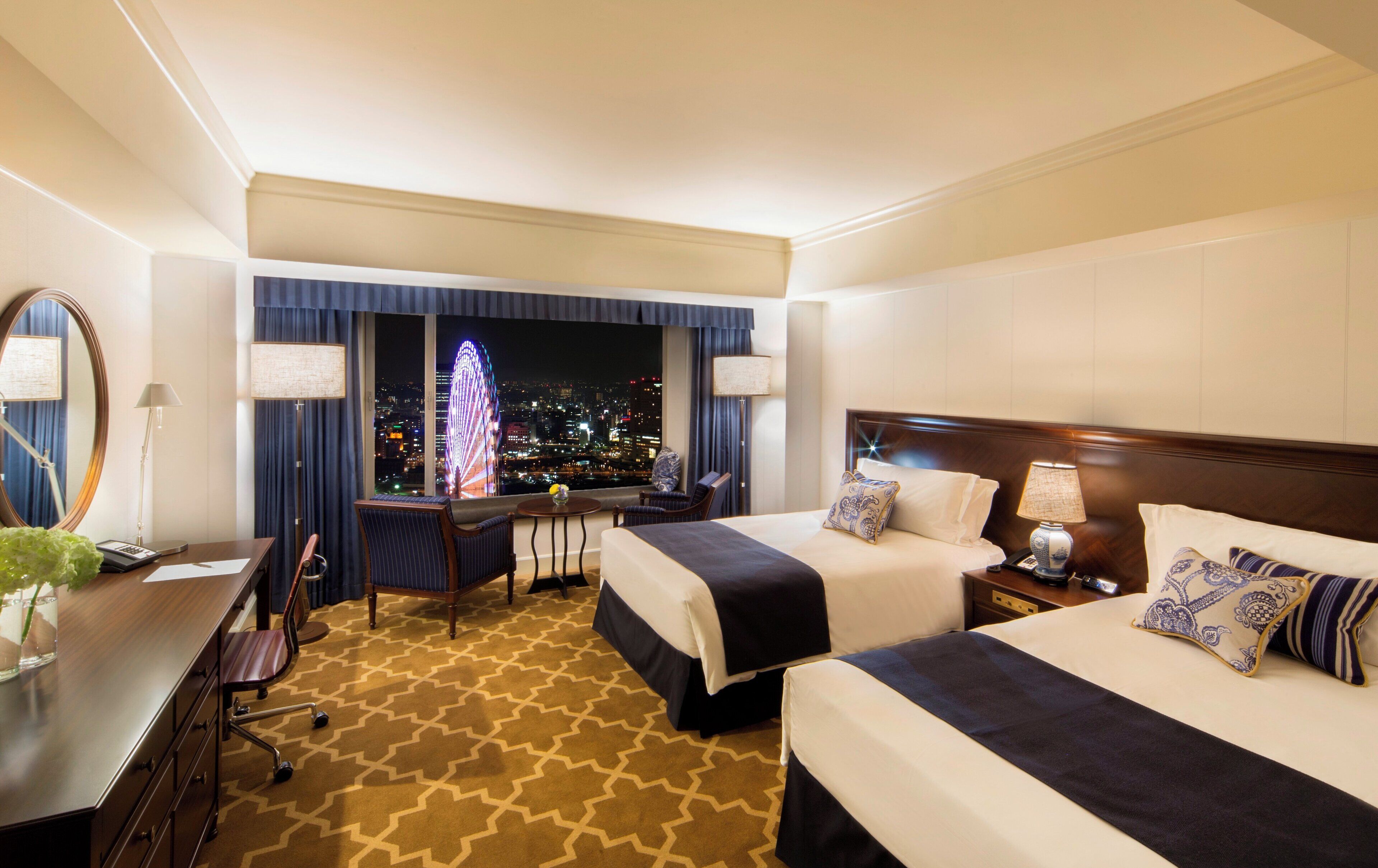 A hotel room with two beds, a desk, a chair, a lamp, and a view of a cityscape at night.