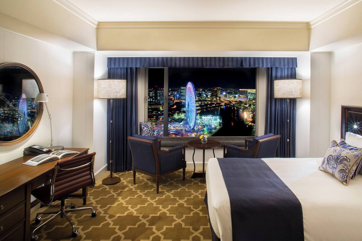 A hotel room with a bed, desk, chair, and a large window with a city view.