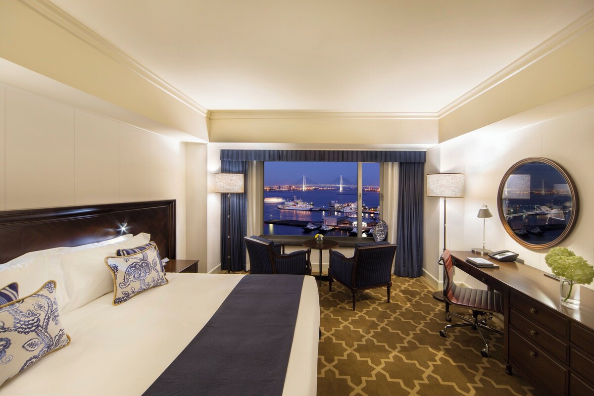 A hotel room with a bed, desk, chair, and a view of a bridge at night.