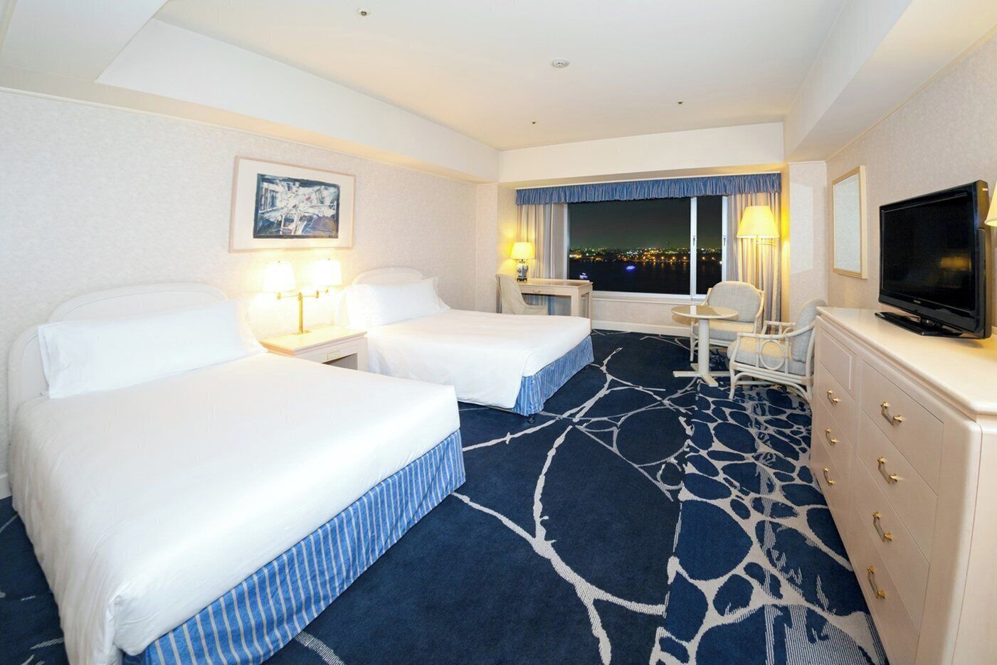 Habitación de hotel con dos camas, un escritorio, un televisor y vista a la ciudad de noche.