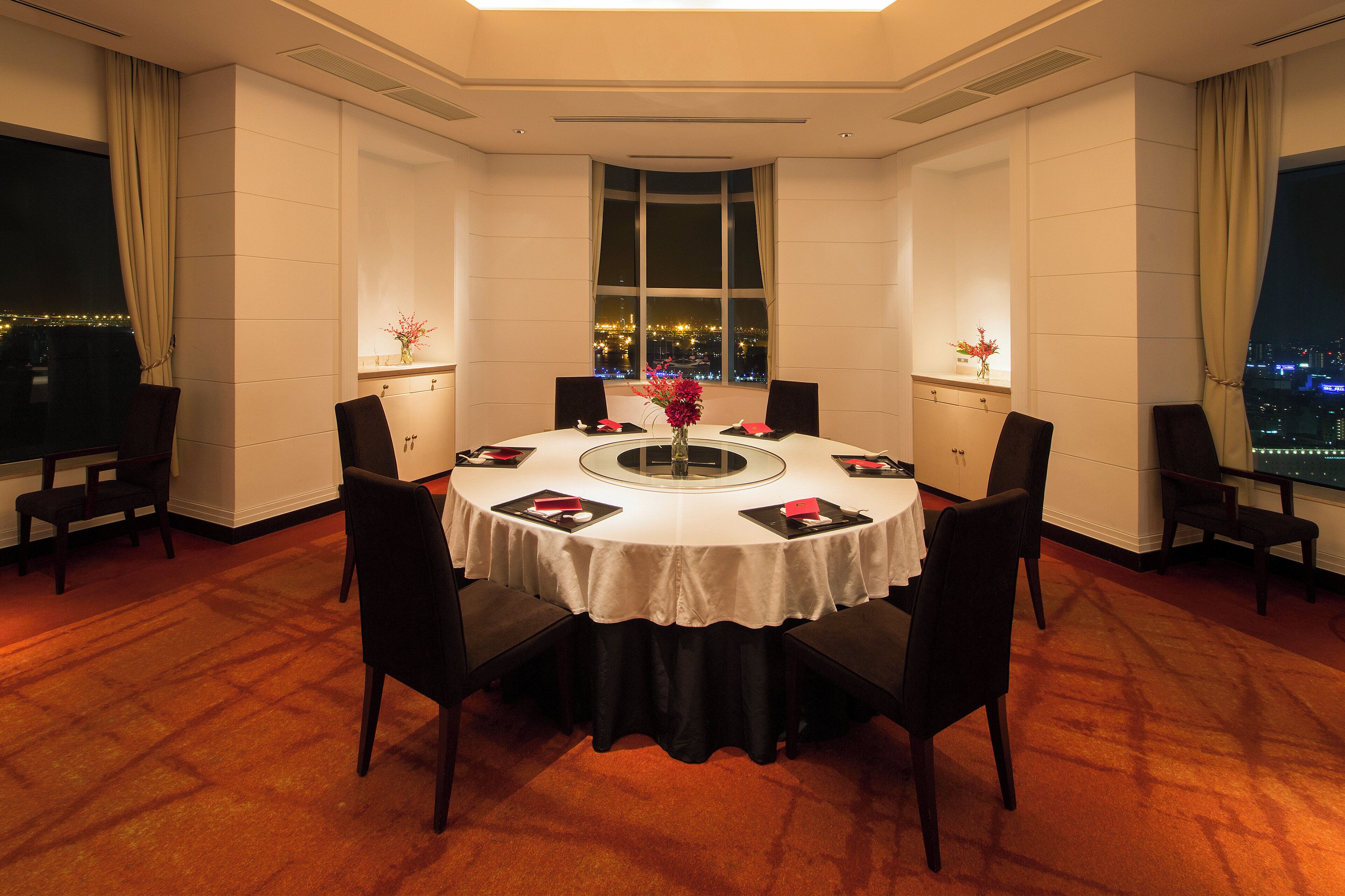 A hotel room with a round dining table, chairs, a vase with flowers, and a view of a cityscape through a large window.