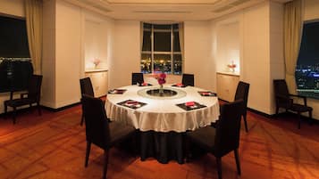 A hotel room with a round dining table, chairs, a vase with flowers, and a view of a cityscape through a large window.