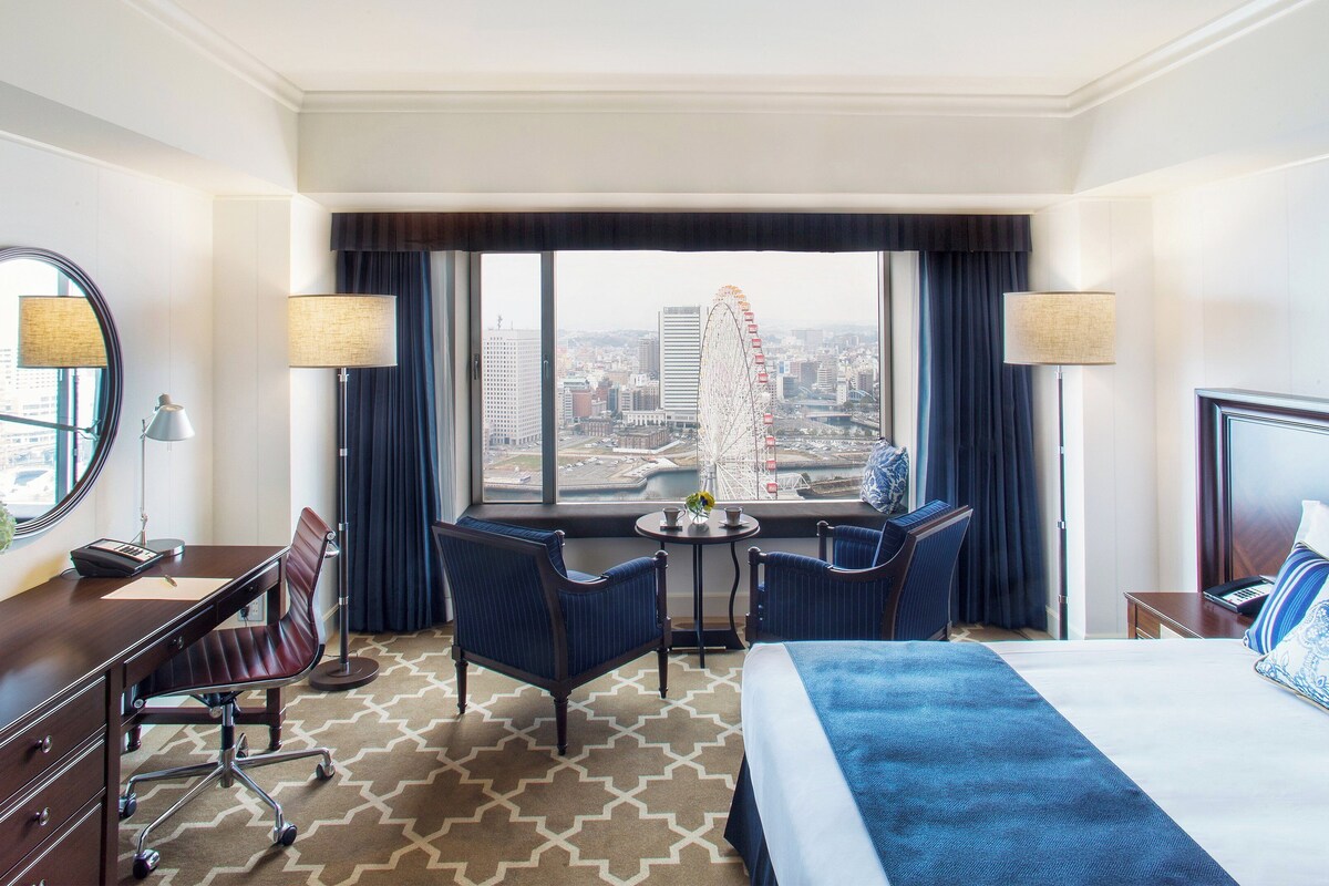 A hotel room with a bed, desk, chair, and a large window with a city view.