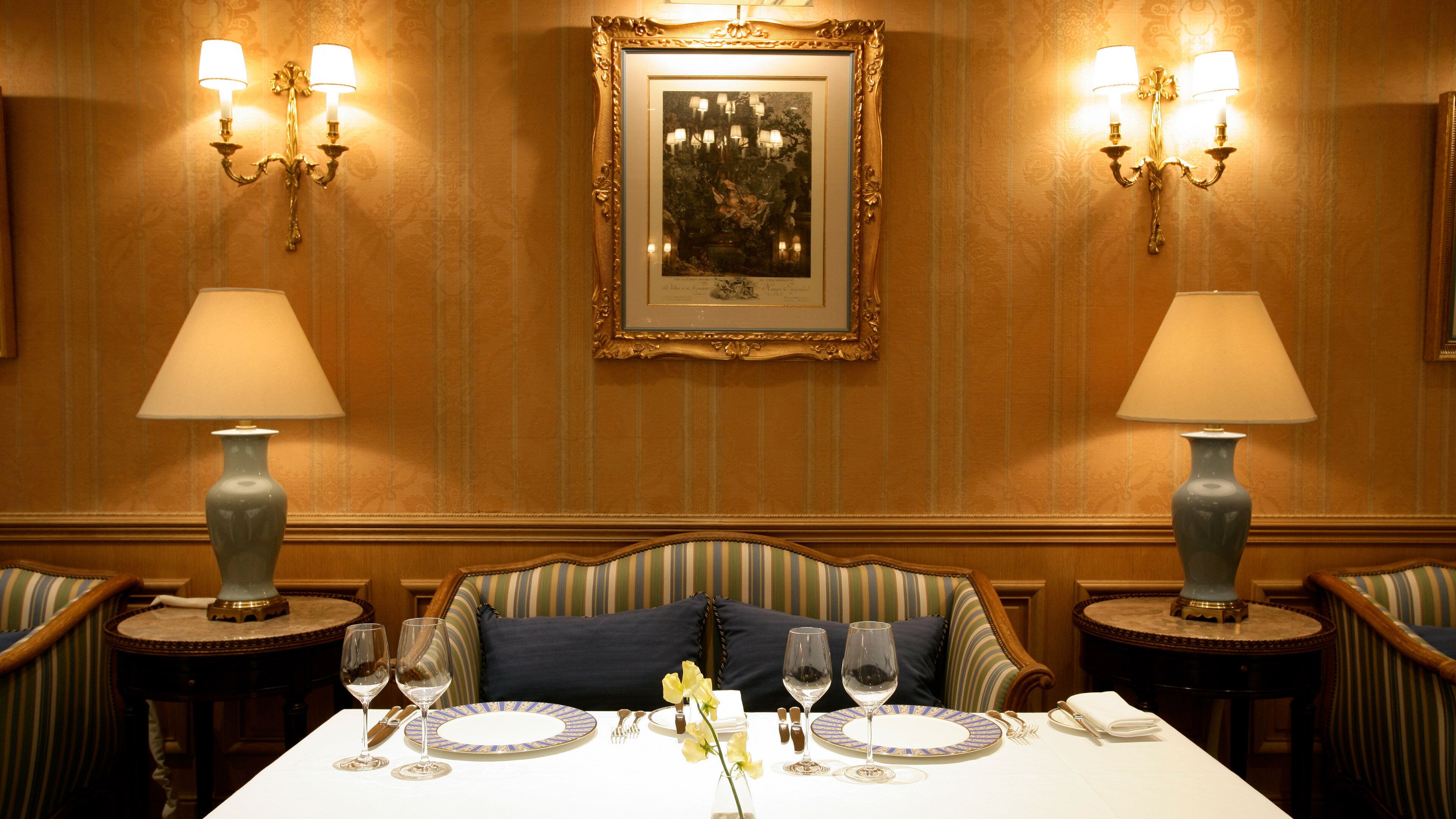 A dining room with a table set for two, flanked by two lamps, a framed artwork on the wall, and a patterned wallpaper.