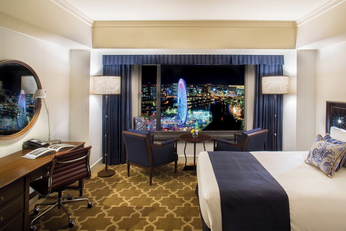 A hotel room with a bed, desk, chair, and a large window with a city view.