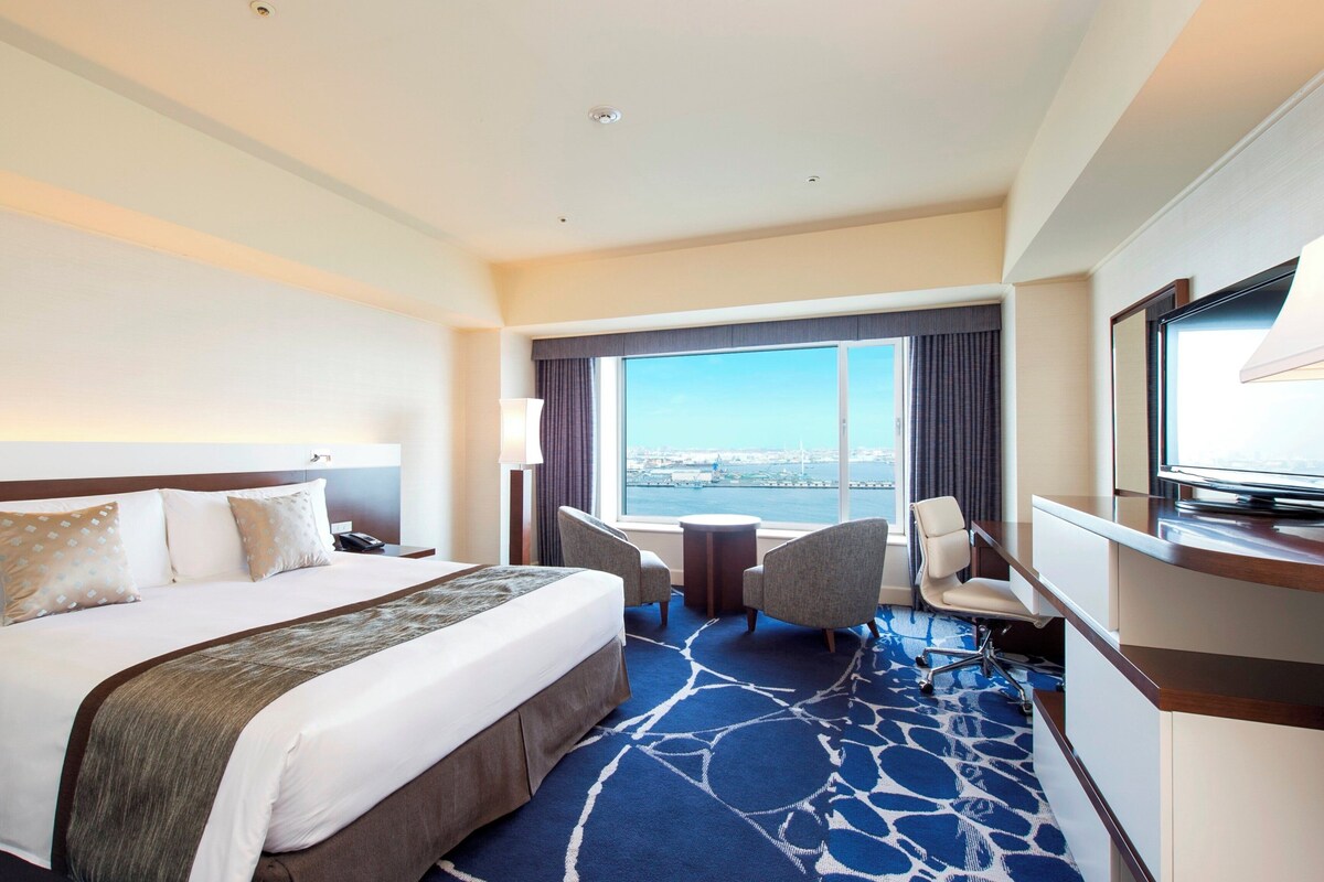 A hotel room with a large bed, a desk with a TV, a chair, and a view of the city.