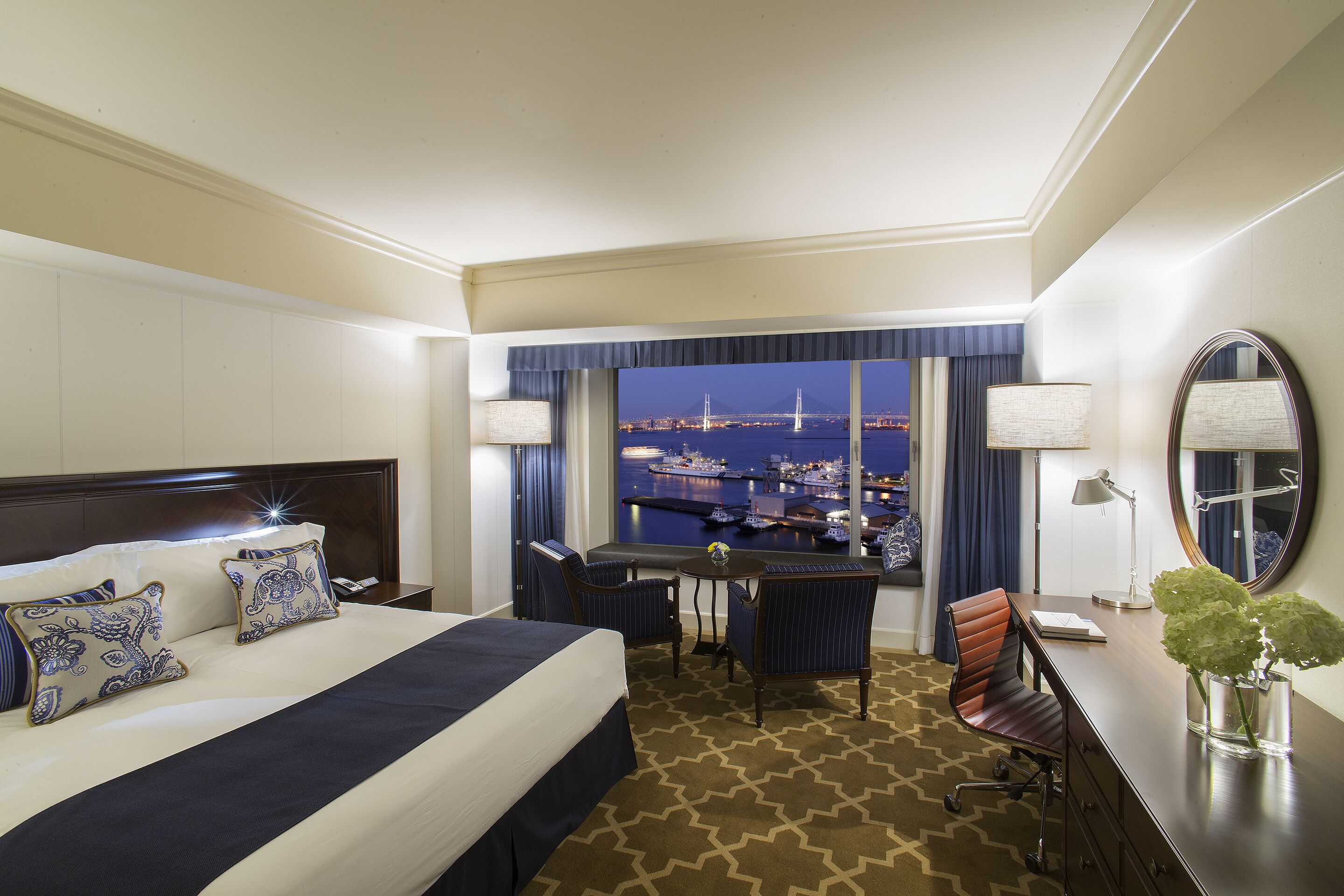Een hotelkamer met een groot bed, een bureau, een stoel en uitzicht op een stadsbeeld 's nachts.