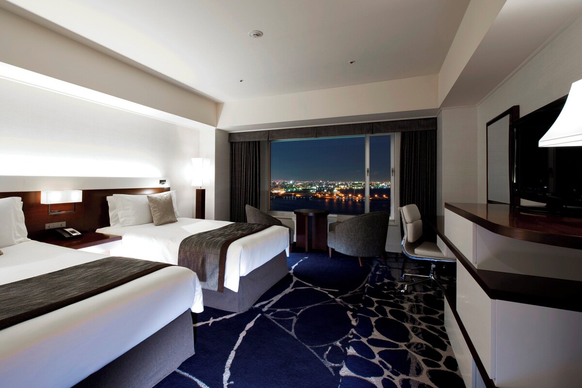 A hotel room with two beds, a desk, a chair, a TV, and a large window with a city view.