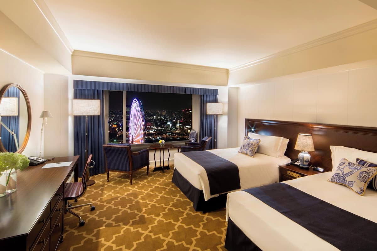 A hotel room with two beds, a desk, a chair, a lamp, and a view of a cityscape at night.
