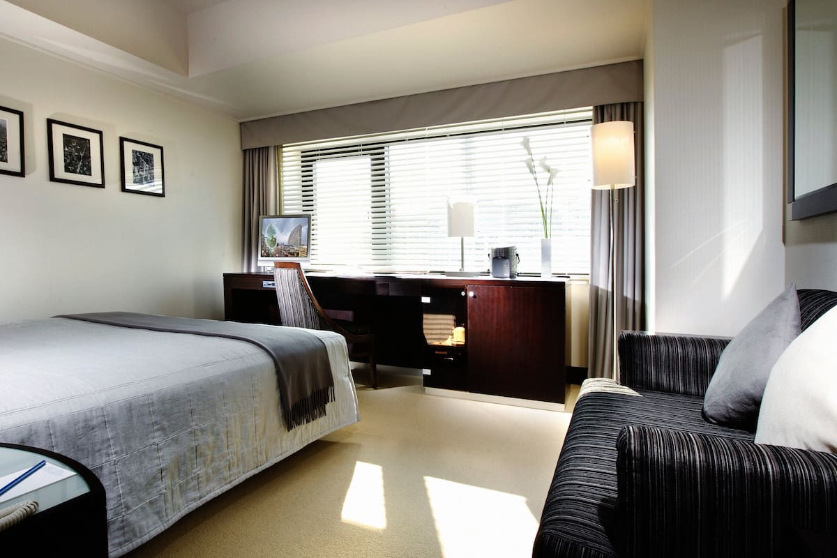 A hotel room with a bed, a desk with a computer, a chair, a sofa, and a window with blinds.