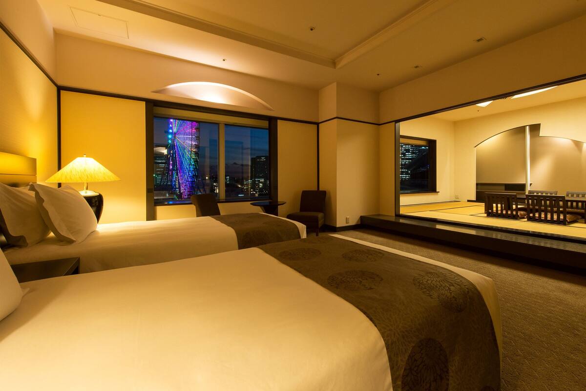 A hotel room with two beds, a sitting area with a table and chairs, a large mirror, and a view of a cityscape at night.