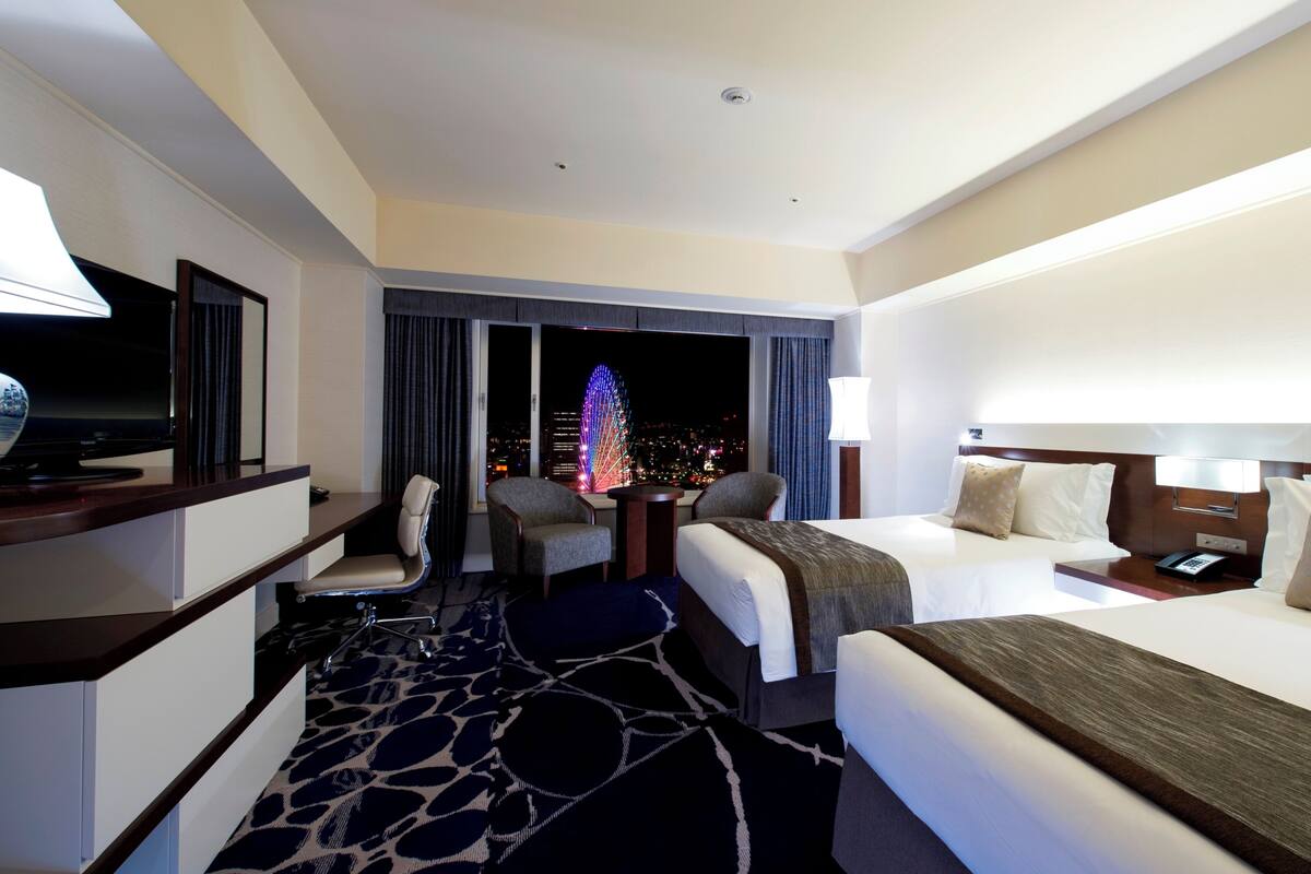 A hotel room with two beds, a desk with a television, a chair, a nightstand, and a view of the city at night.