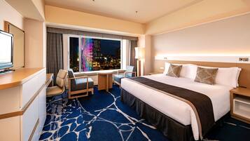 A hotel room with a bed, a desk, a chair, a TV, and a view of a cityscape at night.