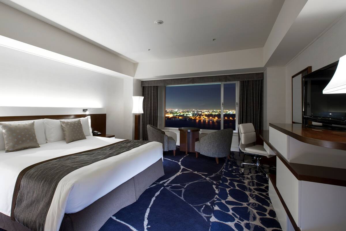 A hotel room with a large bed, a desk, a chair, a TV, and a view of a cityscape at night.