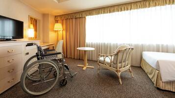 Habitación de hotel con silla de ruedas, una mesa redonda, una silla de mimbre, una cama, un televisor y una lámpara.