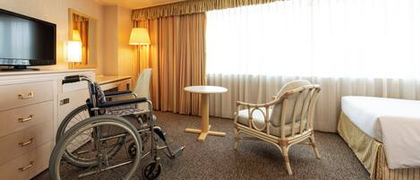 Habitación de hotel con silla de ruedas, una mesa redonda, una silla de mimbre, una cama, un televisor y una lámpara.