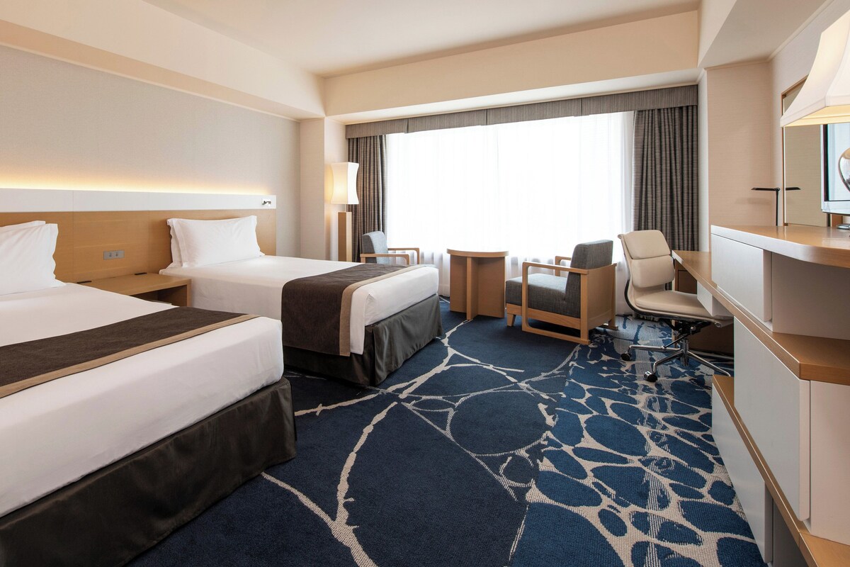 A hotel room with two beds, a desk, a chair, and a small table.