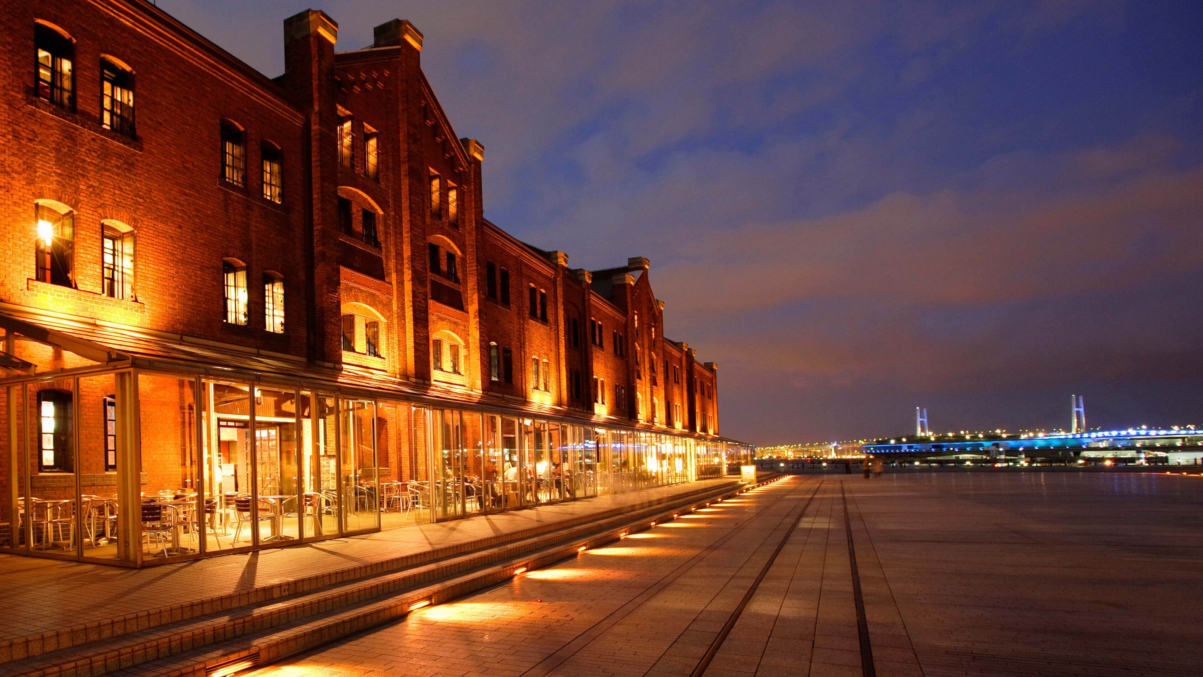 A waterfront building with large windows and a brick facade, overlooking a river with a bridge in the distance.