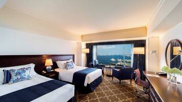 Quarto de hotel com duas camas, uma mesa, uma cadeira, uma janela grande com vista para o mar e um espelho.