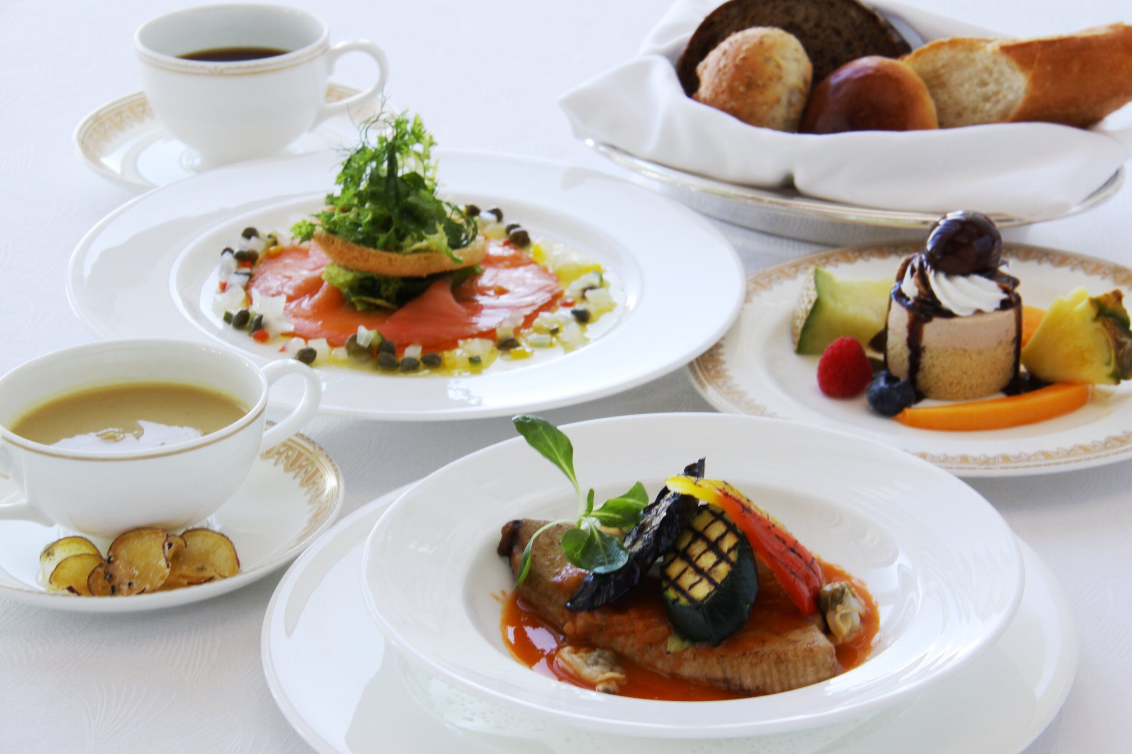 Una mesa con varios platos, incluyendo un plato con verdura a la parrilla y pescado, un postre con bayas, y un bol de pan.