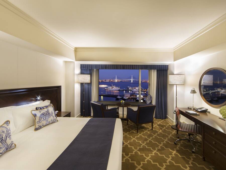 Quarto de hotel com cama, escrivaninha, cadeira e vista noturna de uma ponte.