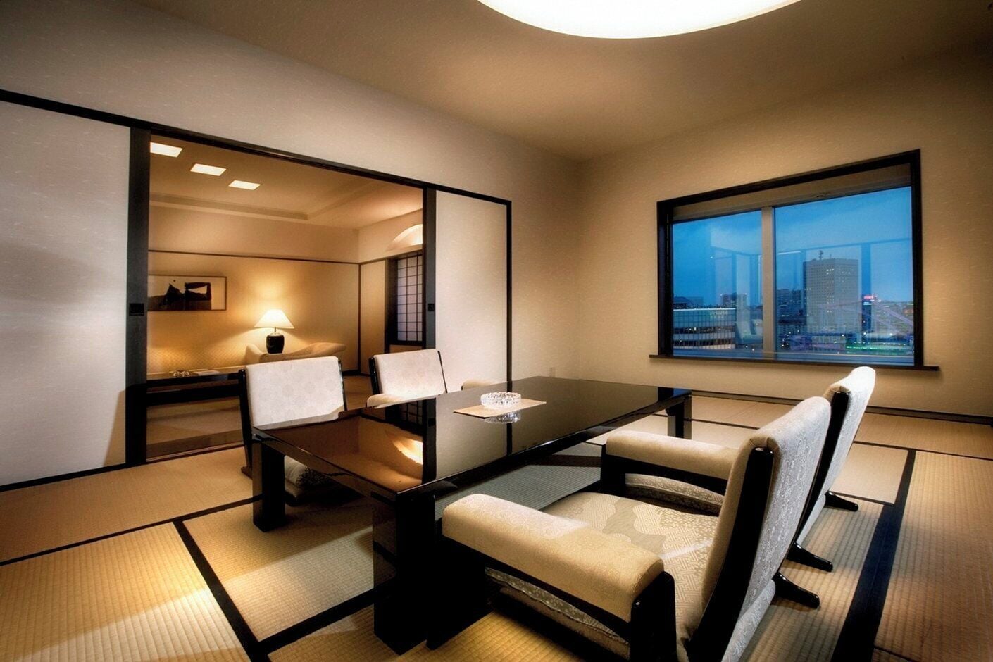 Habitació d'hotel moderna amb taula de menjador, cadires, un sofà, una finestra amb vista a la ciutat i esteves al terra.