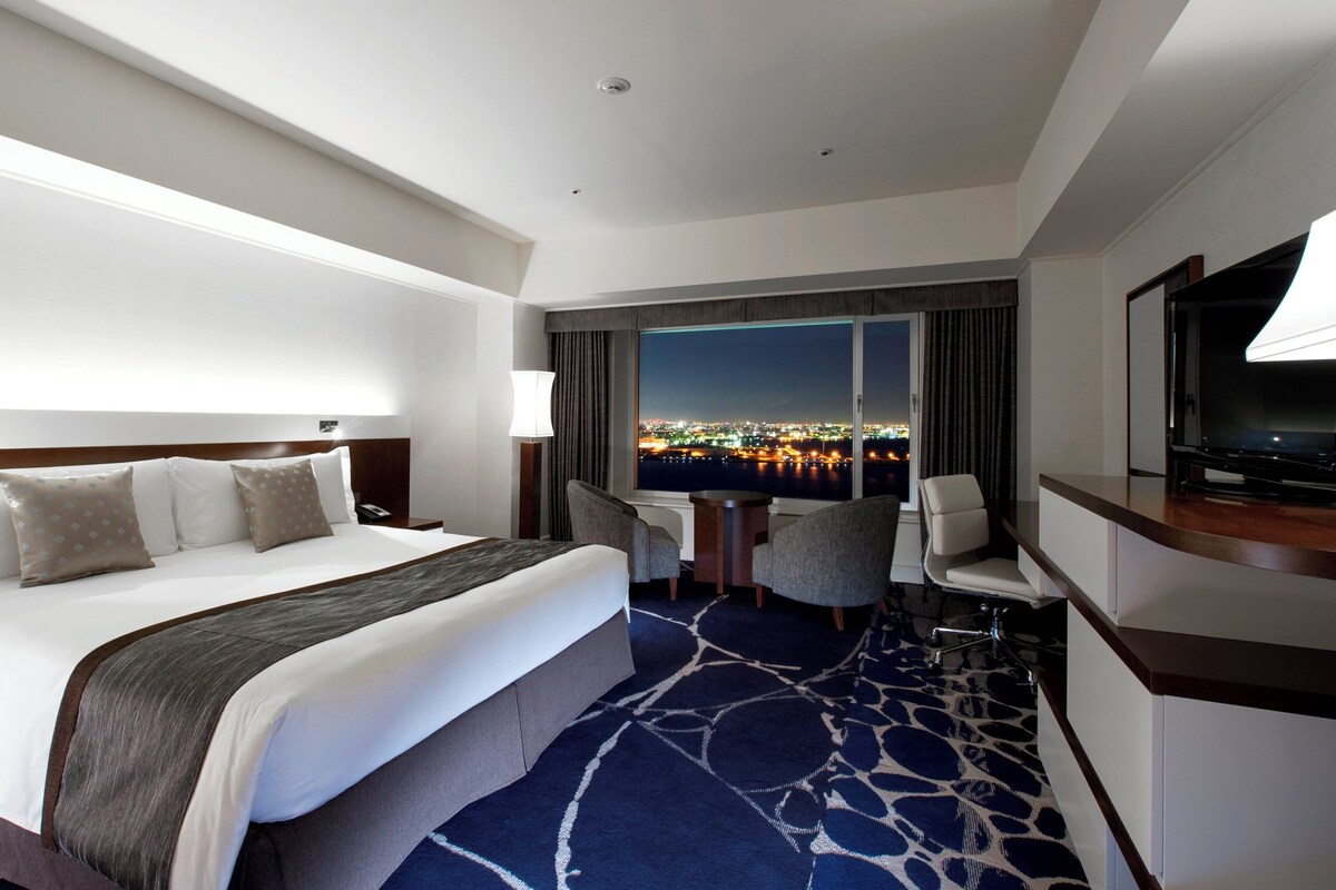 A hotel room with a bed, a desk, a chair, a TV, and a large window with a city view.