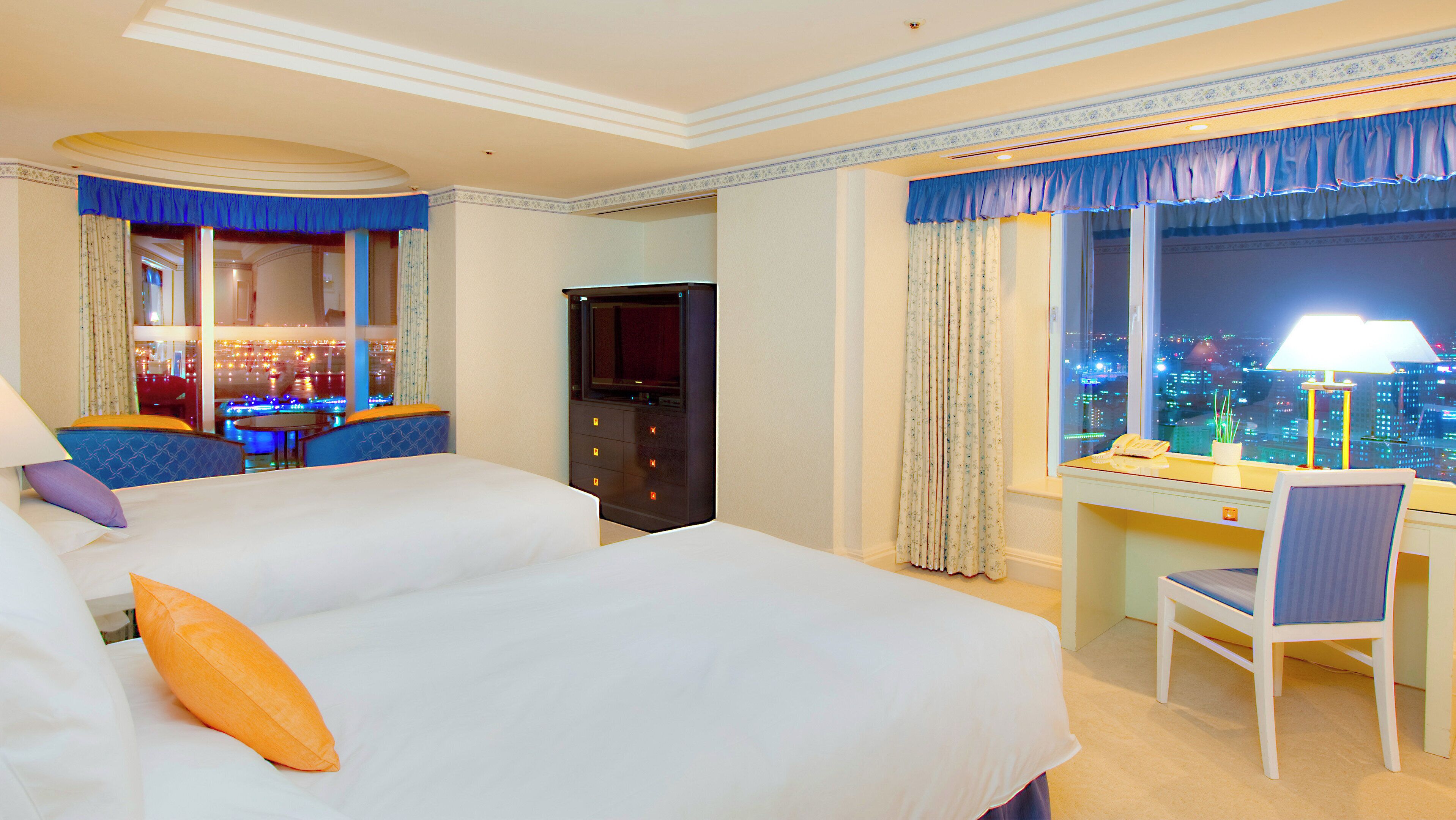 Habitación de hotel con dos camas, un escritorio, una silla, un televisor y vistas a la ciudad.