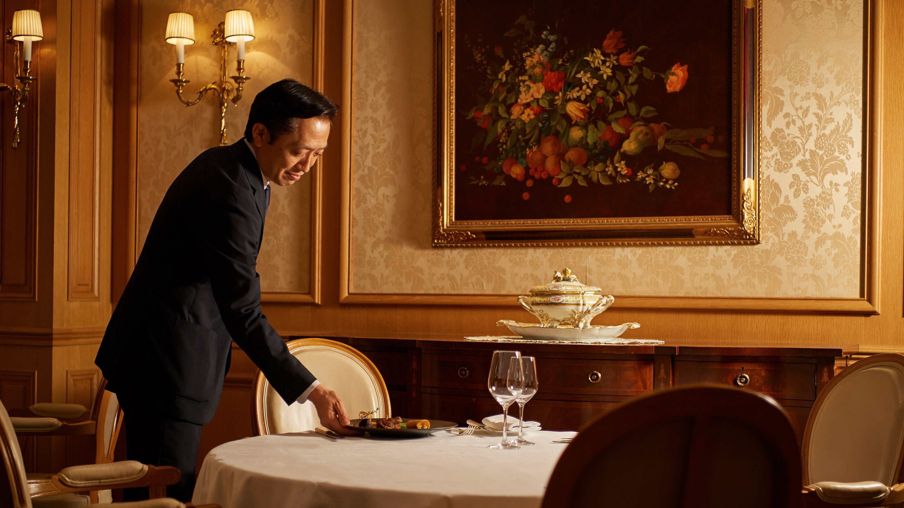 A man serving food at a table in a room with a floral painting and a gold-framed mirror.