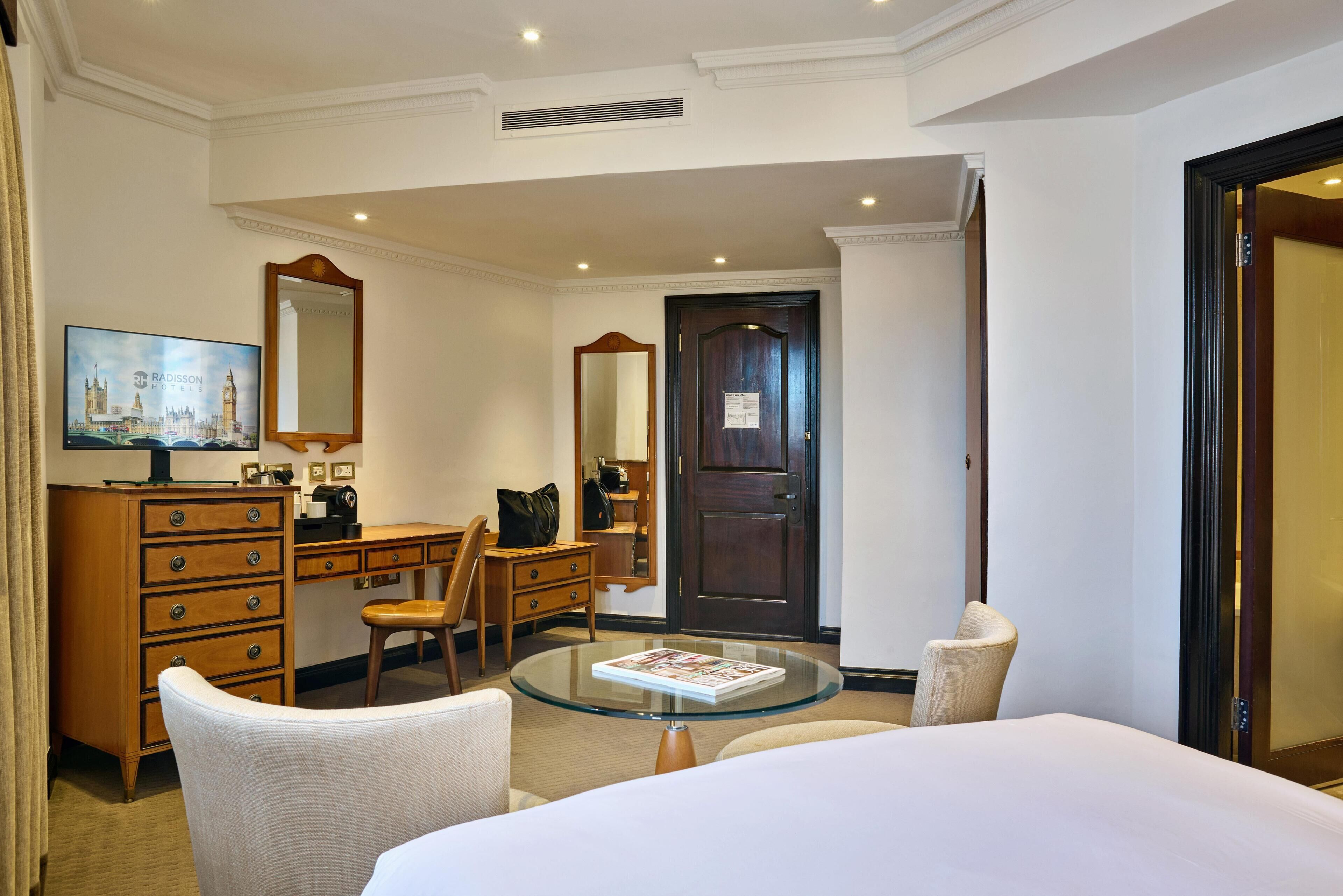 A hotel room with a bed, a glass-top table, a wooden dresser, a desk, a chair, a mirror, and a door.