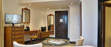 A hotel room with a bed, a glass-top table, a wooden dresser, a desk, a chair, a mirror, and a door.