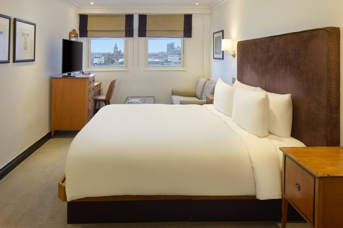 A hotel room with a large bed, a desk with a TV, a chair, and a view of the city through the windows.