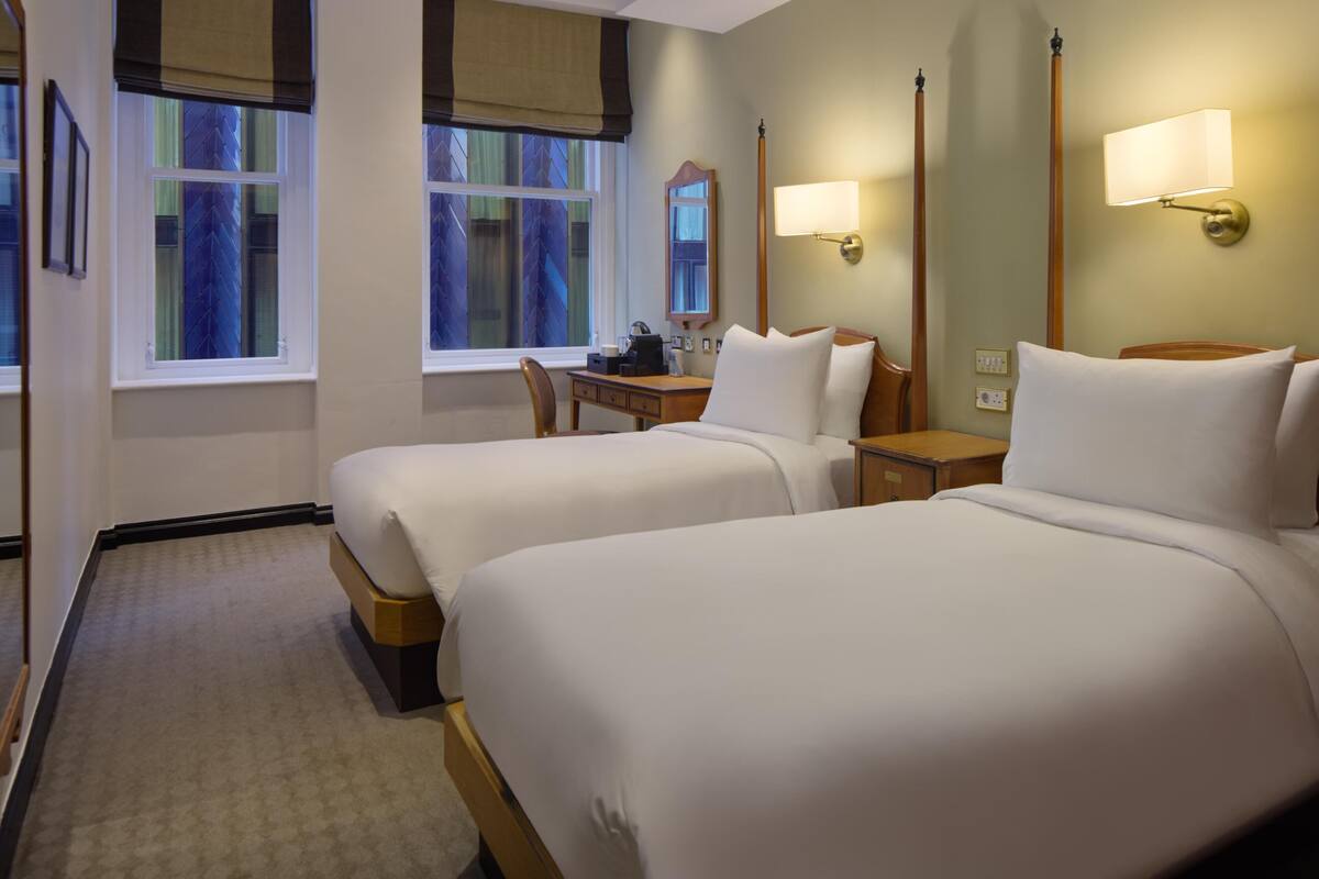 A hotel room with two beds, a desk, a chair, and a window with a view of trees.