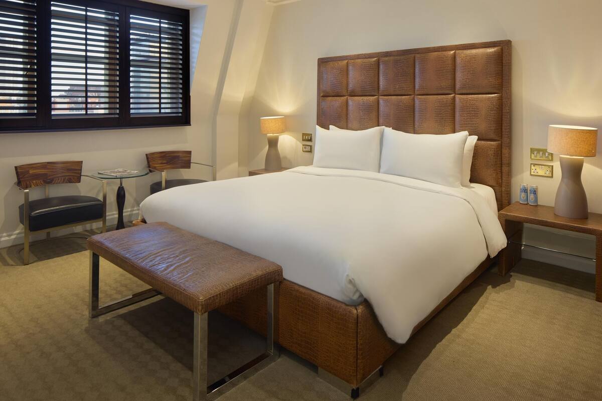A hotel room with a large bed, two bedside tables with lamps, a bench, a small table, and chairs.