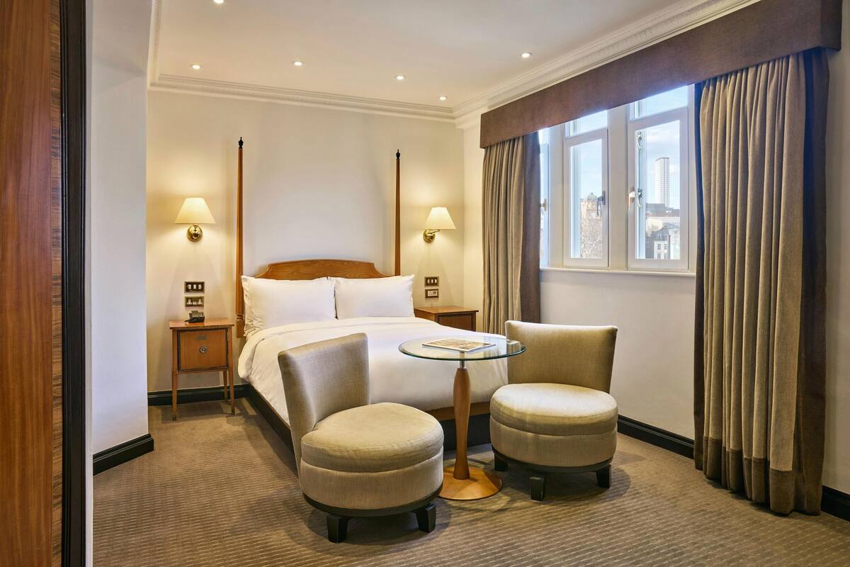 A hotel room with a bed, two armchairs, a small table, two bedside lamps, a window with curtains, and a view of buildings.