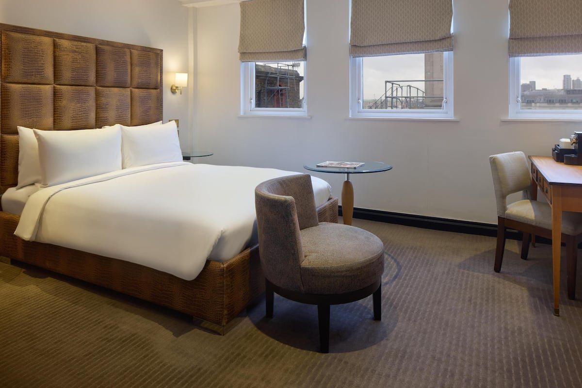 A hotel room with a large bed, two chairs, a small table, and a view of the city through the windows.