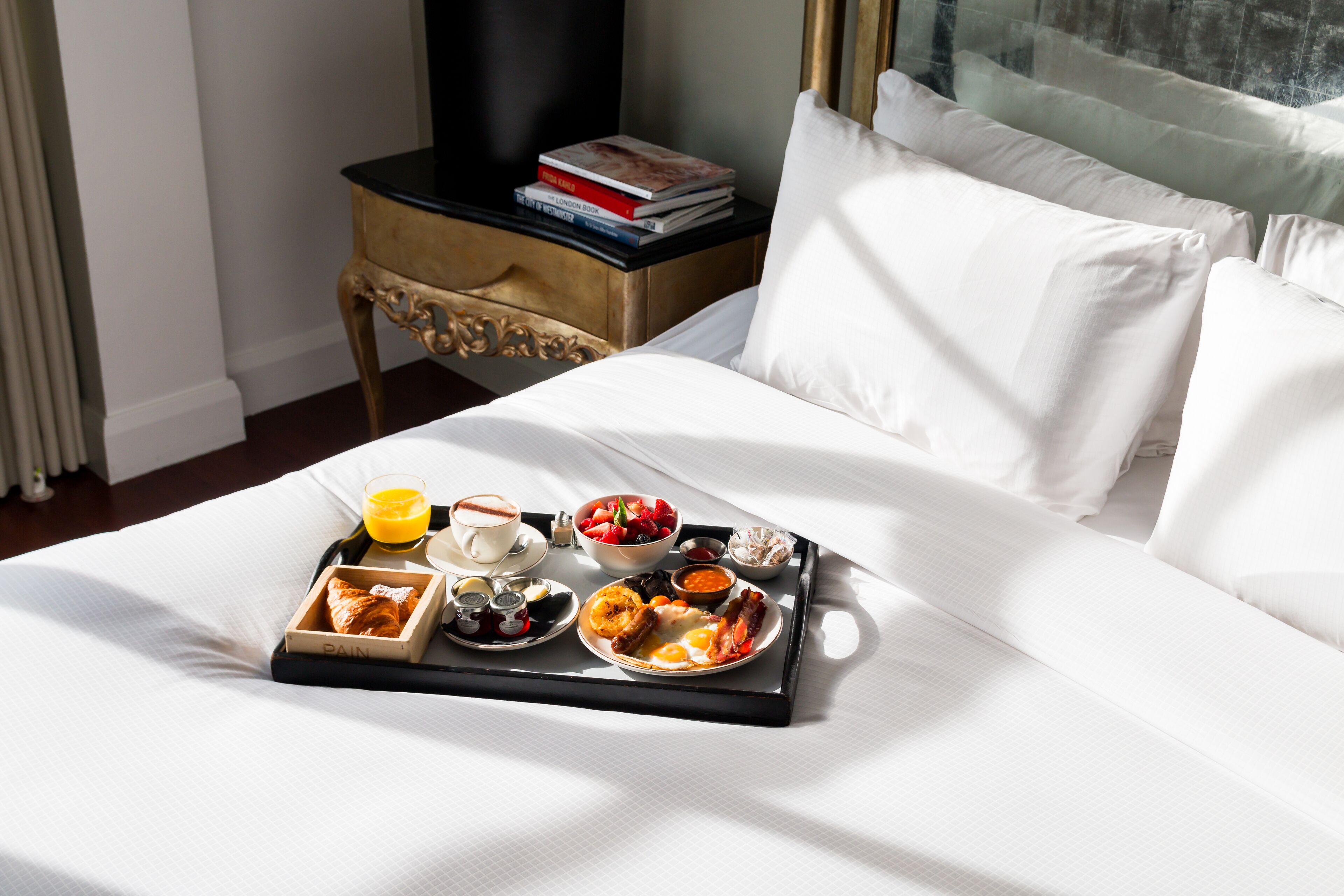 A neatly made bed with a tray of breakfast including eggs, toast, and fruit, a bedside table with books, and a window with curtains.