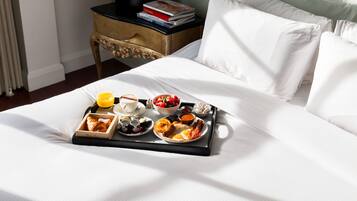 A neatly made bed with a tray of breakfast including eggs, toast, and fruit, a bedside table with books, and a window with curtains.