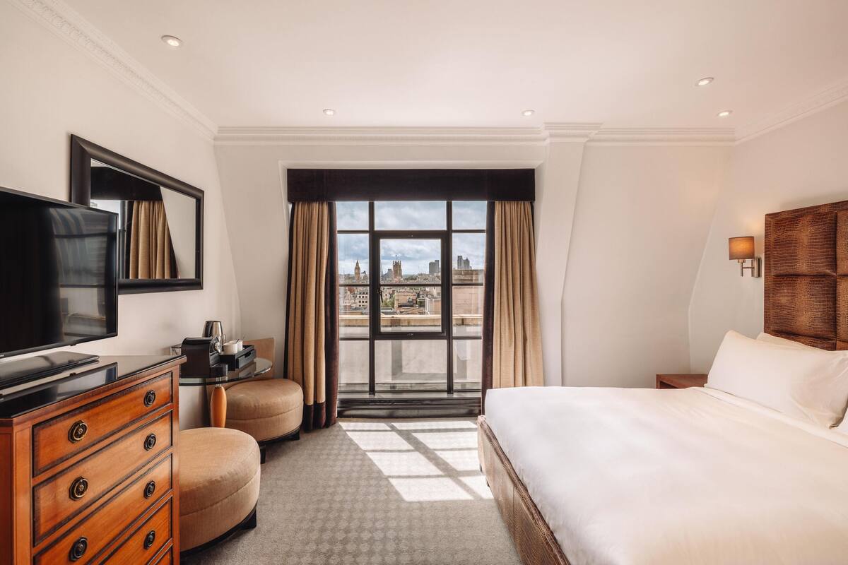 A hotel room with a bed, a television, a dresser, a chair, a desk, and a window with a city view.