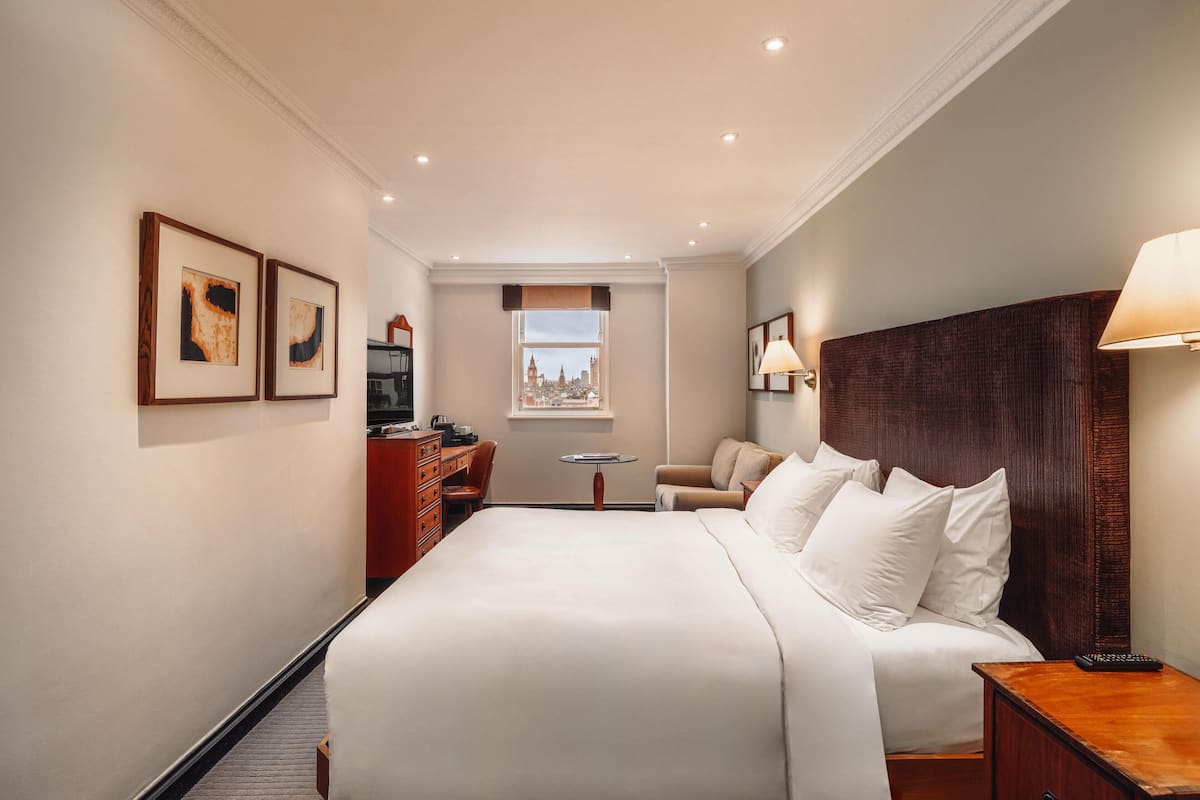 A hotel room with a large bed, a desk, a chair, a small table, and a window with a city view.