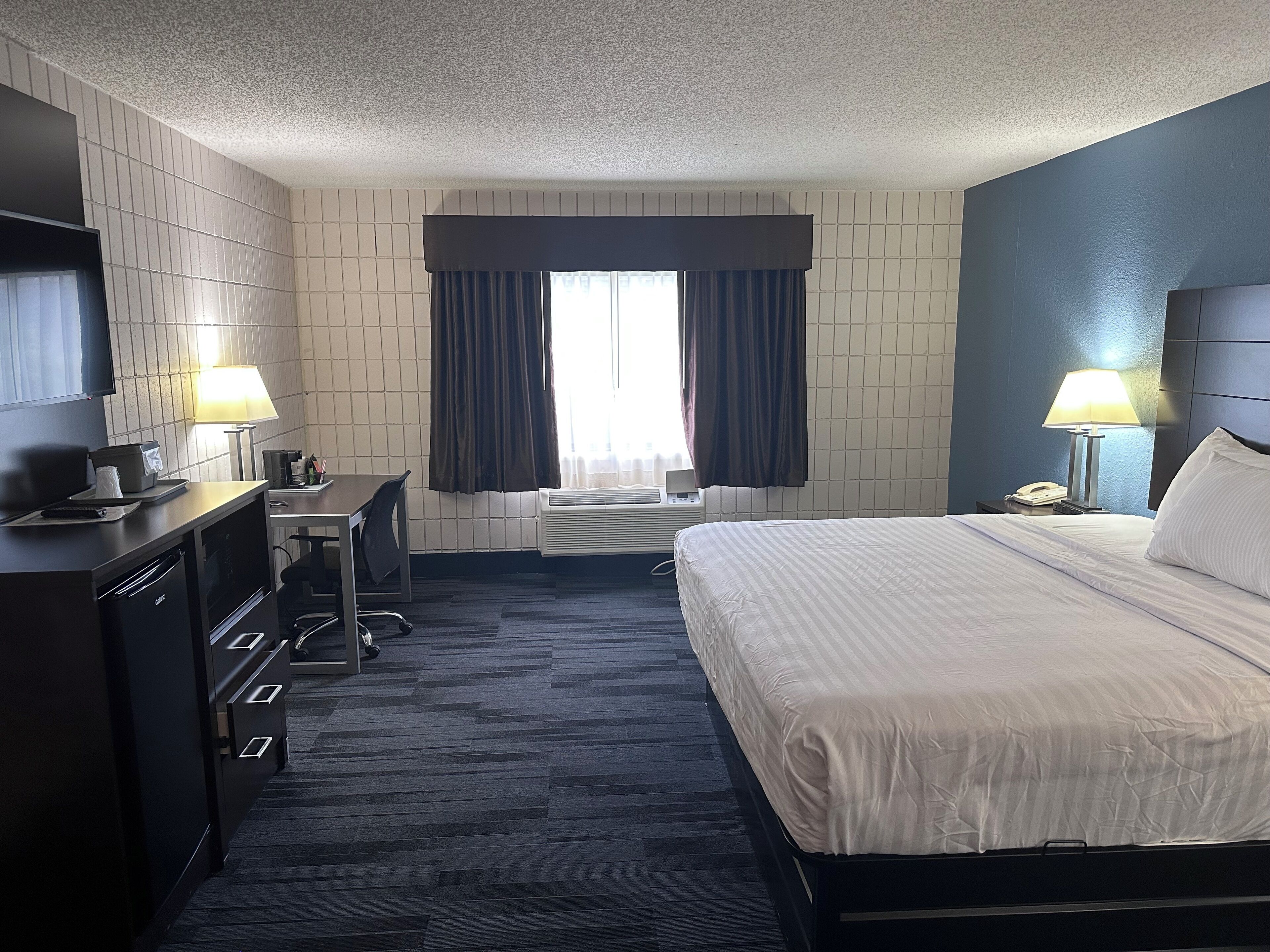 A hotel room with a bed, desk, chair, TV, lamps, and a window with curtains.